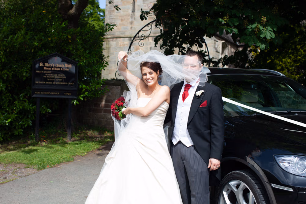 Bride and groom laughing as the wind lifts her veil beside their wedding car at St Mary’s Church in Wath. Captured naturally by Tim Christian Jones