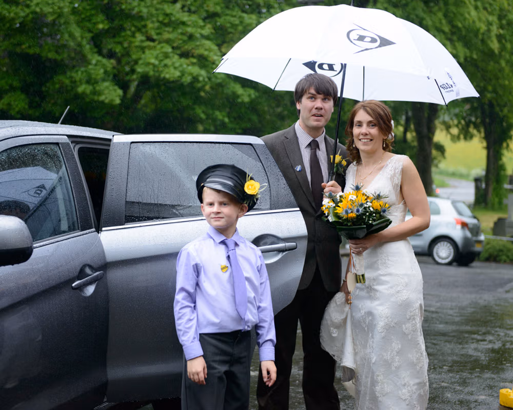 Wedding couple standing under a white umbrella beside their car at Barley Village Hall, their son wearing a driver’s cap as rain gathers overhead.