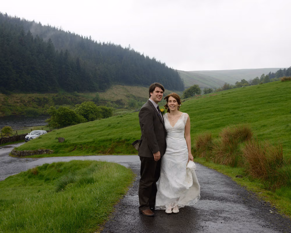 Wedding couple standing together on a quiet country road on Pendle Hill, with green banks, trees and hills stretching away in soft rain.