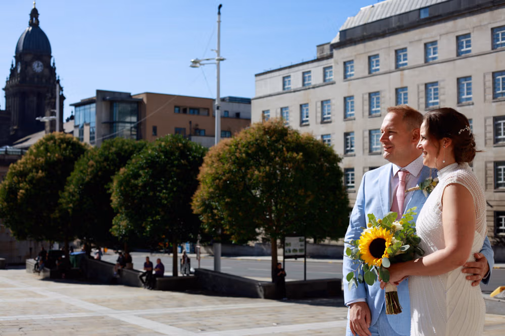 Gemma and Danny standing together on the steps of Leeds Civic Hall, smiling across Millennium Square in sunshine.