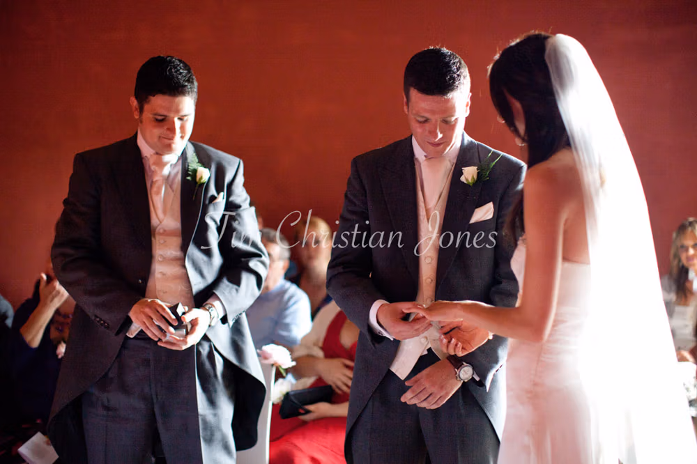 Groom places the wedding ring on the bride’s finger as best man holds the ring box