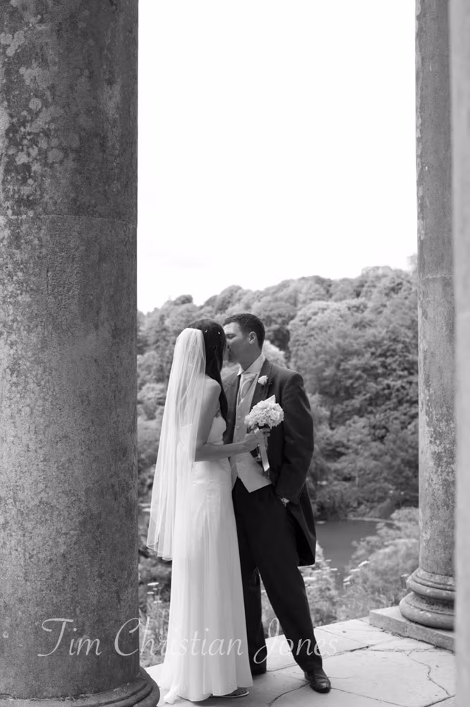 Bride and groom kiss beneath stone arches with trees behind them